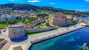 A coastal town with a prominent historical fortification, a paved promenade, and a clear view of the sea.