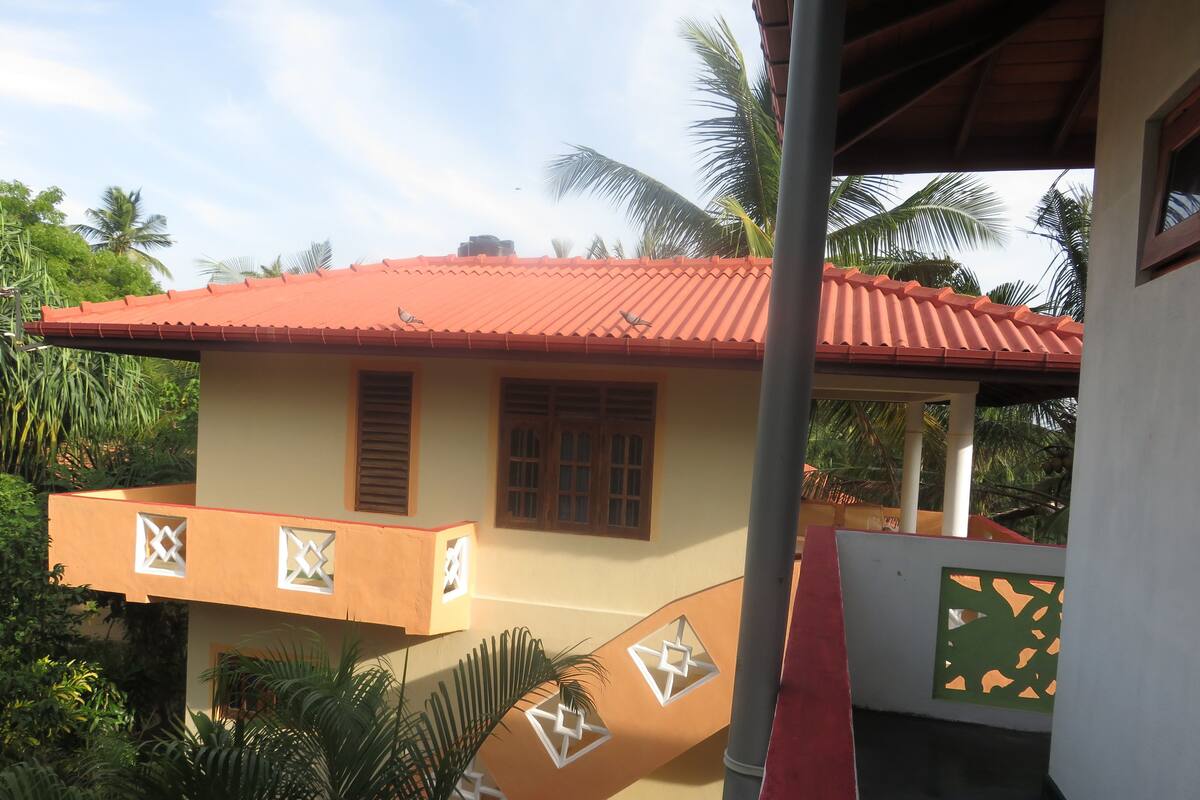 A two-story building with a red-tiled roof, a balcony with geometric patterns, and a surrounding lush garden.