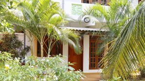 Um edifício tradicional com telhado ladrilhado, portas de madeira e um pequeno alpendre rodeado por vegetação exuberante e palmeiras.