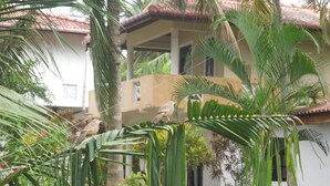 Uma casa com telhado de cerâmica, cercada por vegetação exuberante, incluindo palmeiras e outras plantas.