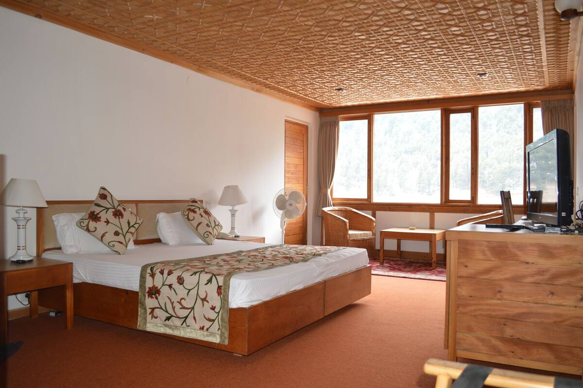 A hotel room with a wooden bed, a desk, a chair, a TV, and a large window.