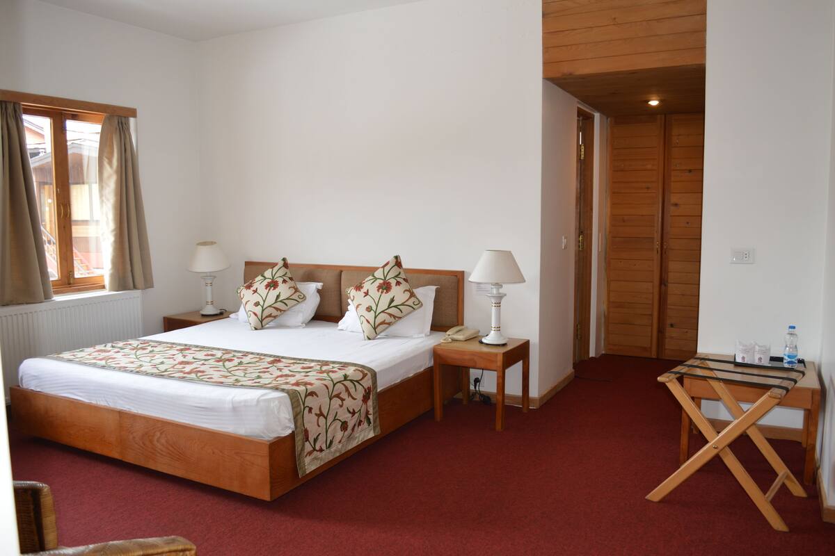 A hotel room with a wooden bed, two bedside tables with lamps, a red carpet, a wooden folding table, and a wooden wardrobe.