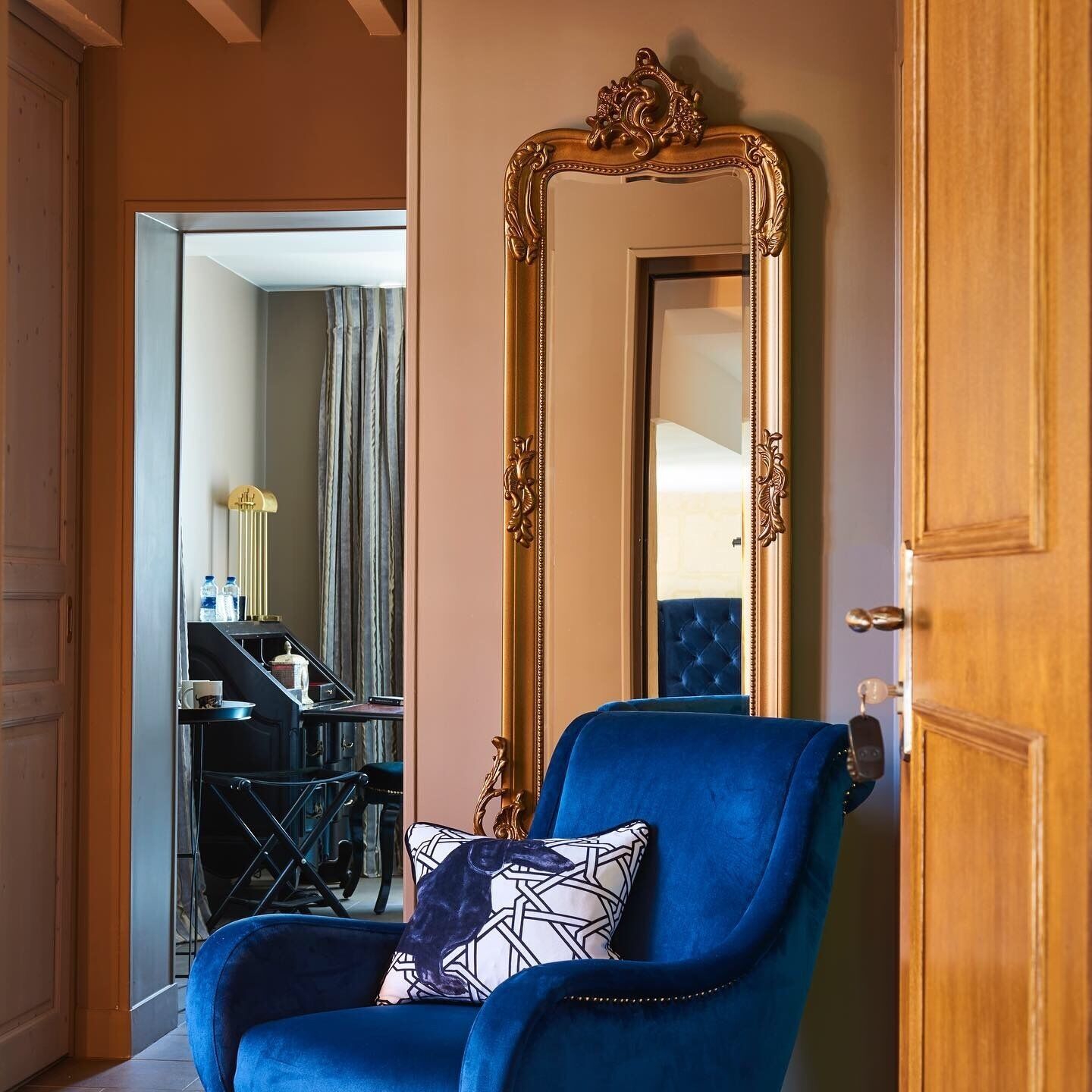 A room with a blue armchair, a large ornate mirror, and a view into another room with a piano and curtains.