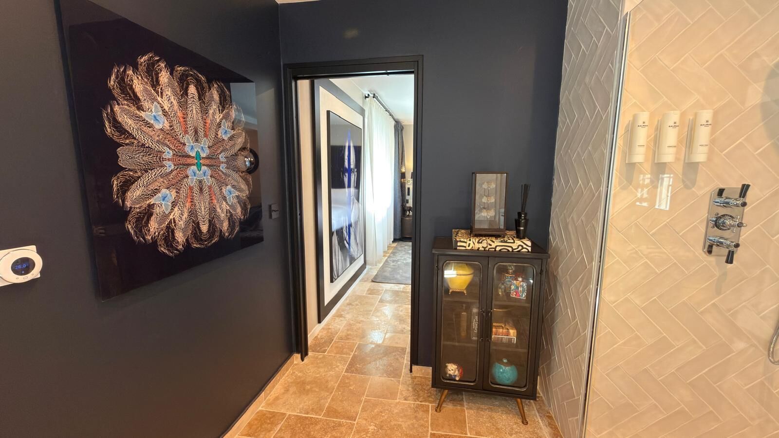 A hallway with a peacock feather artwork, a cabinet with toiletries, and a tiled floor.