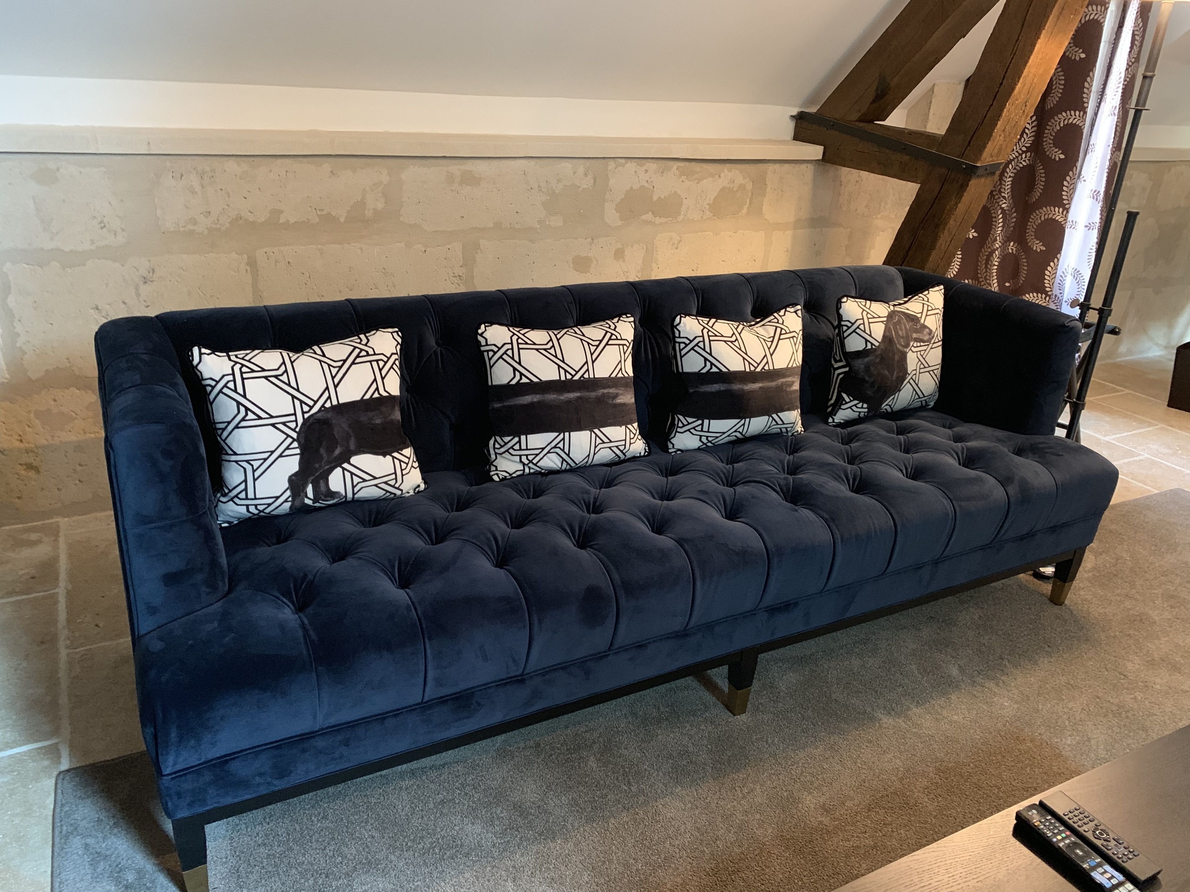 Un sofá azul oscuro con capitoné y cojines estampados, ubicado en una habitación con paredes de ladrillo visto y vigas de madera.