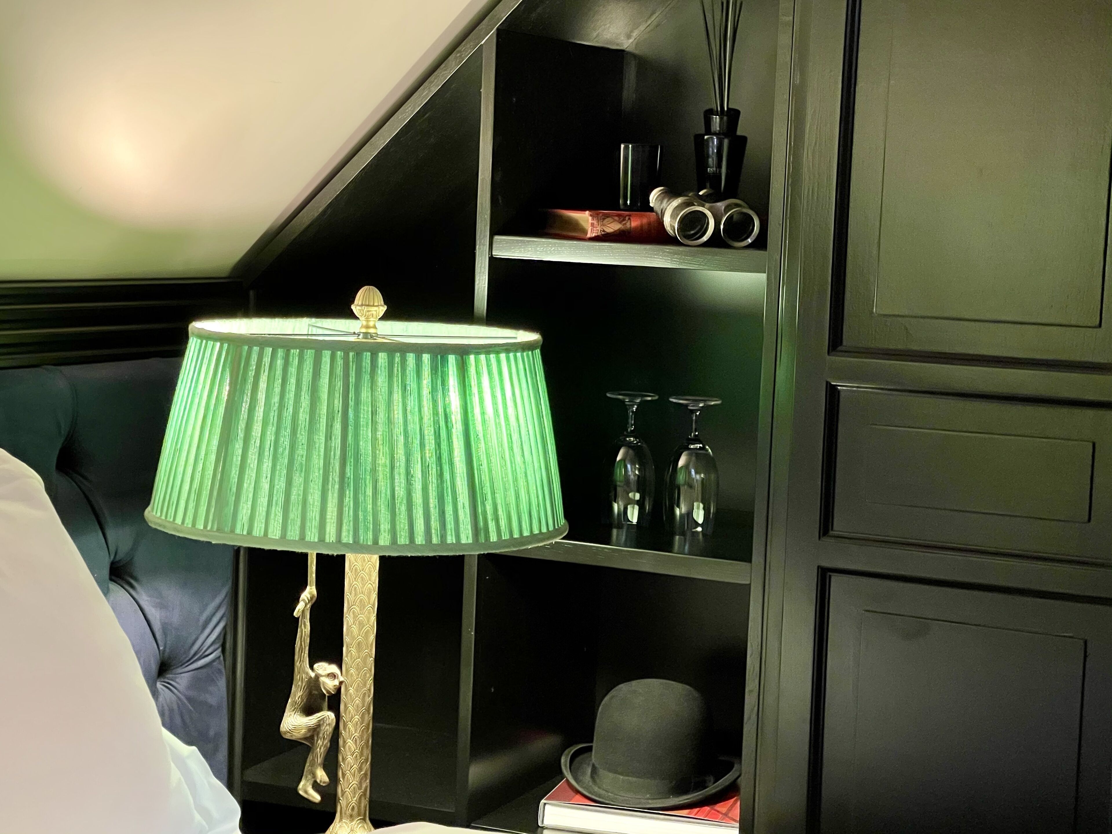 A room with a green lamp, a bookshelf with books and a hat, and a dark-colored bed.