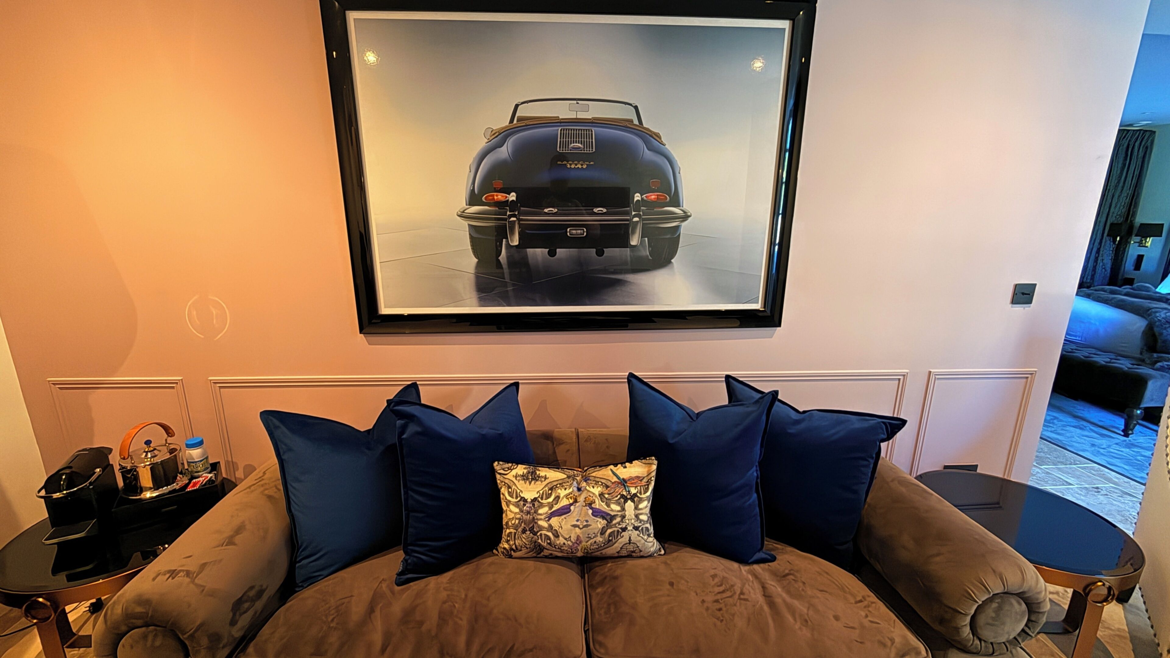 Une pièce avec un canapé marron, un tableau encadré représentant une voiture et une petite table sur laquelle se trouvent une bouilloire et une bouteille.