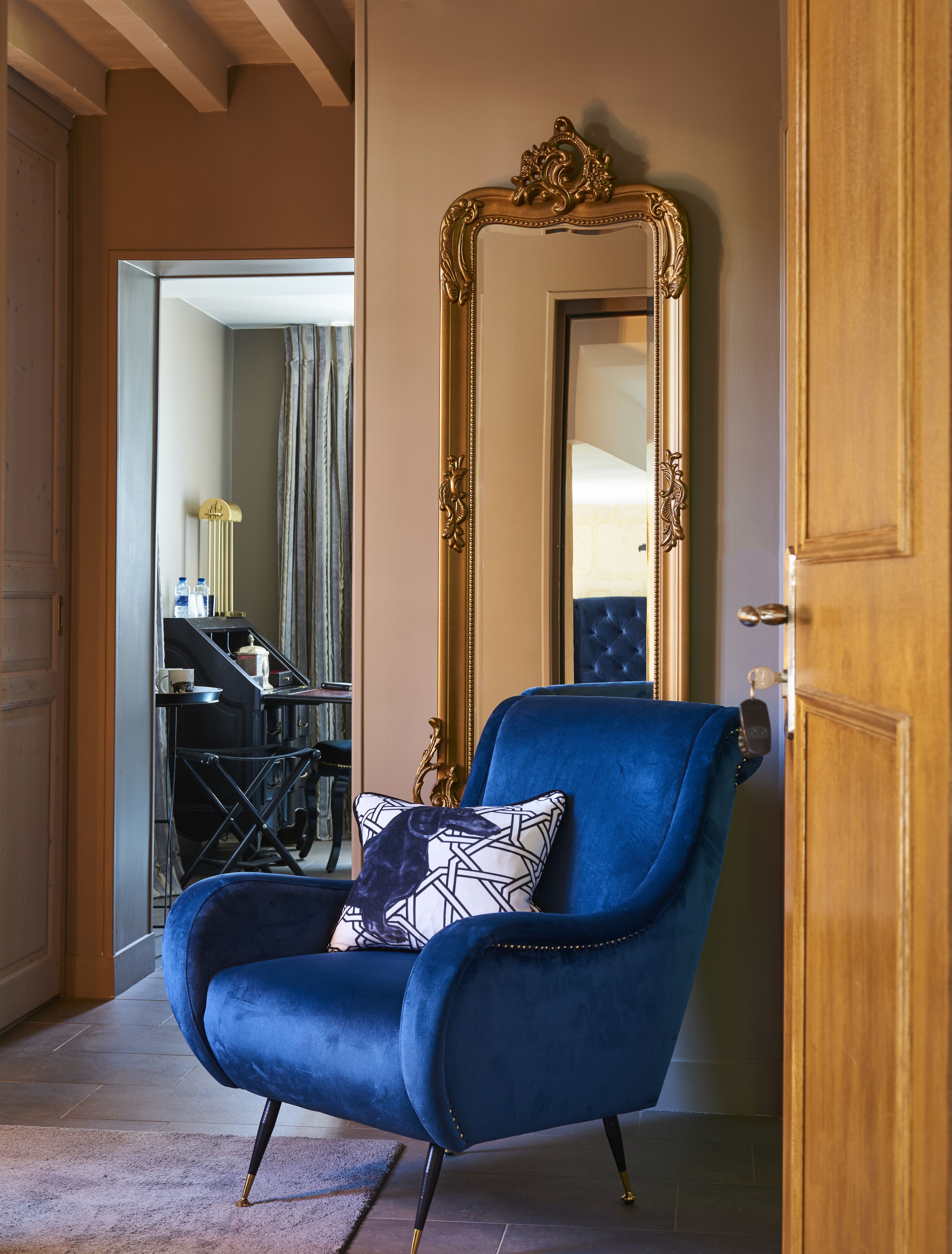 A blue armchair with a patterned pillow in a room with a large ornate mirror and a wooden door.