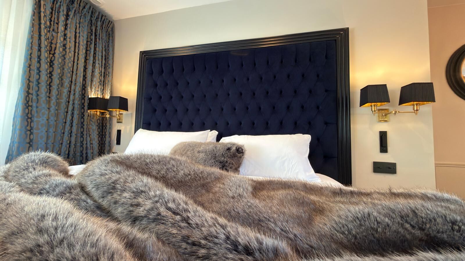 Ein Bett mit dunkelbrauner Kopfteil, weißen Kissen und einem Fellüberwurf. An beiden Seiten des Bettes befinden sich Wandlampen.