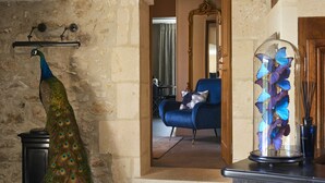 Un pavo real en una habitación con paredes de piedra, una silla azul y una cúpula de vidrio con mariposas.