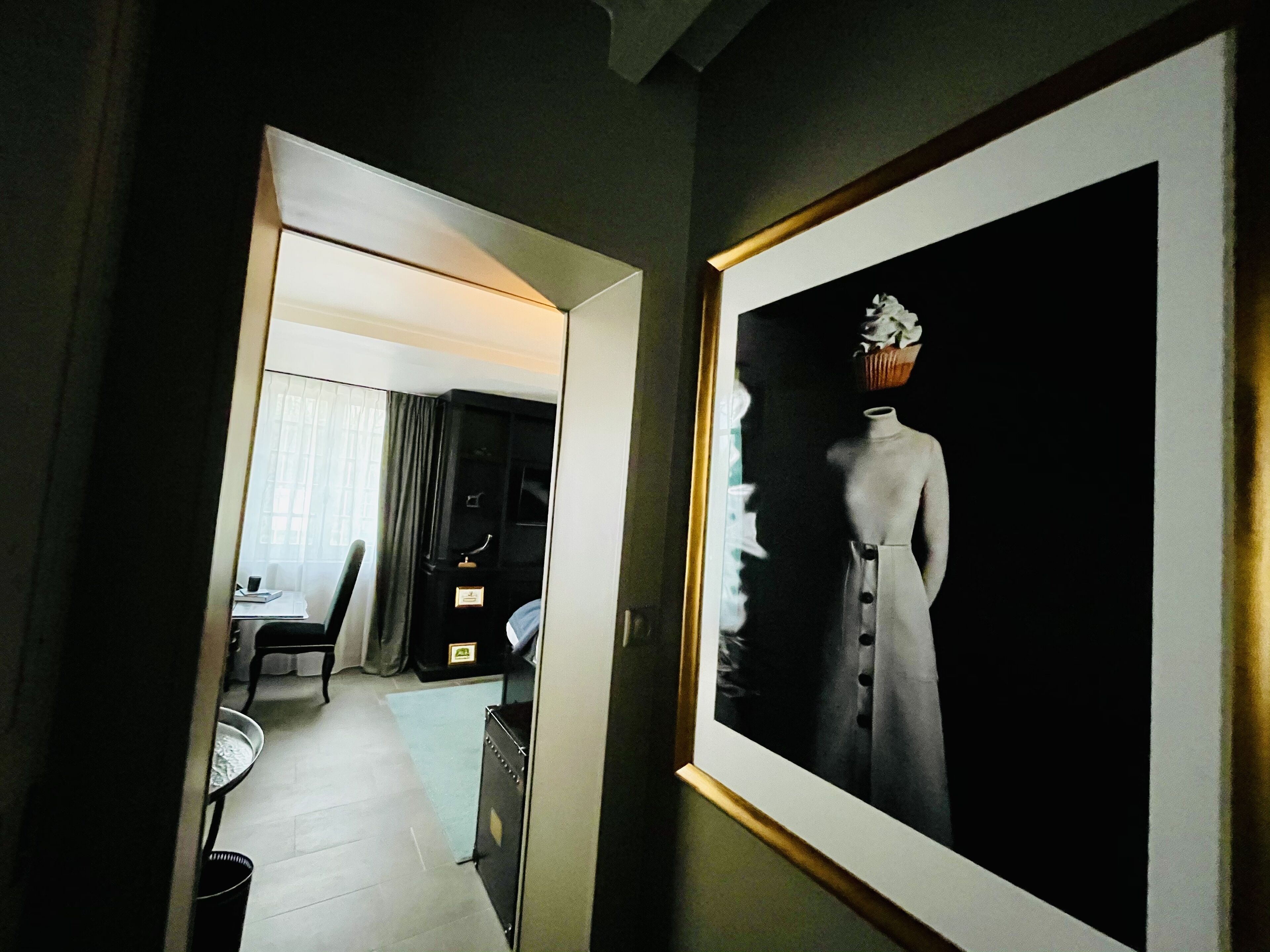 Un pasillo con una obra enmarcada de un maniquí vestido, un escritorio con una silla y una ventana con cortinas.