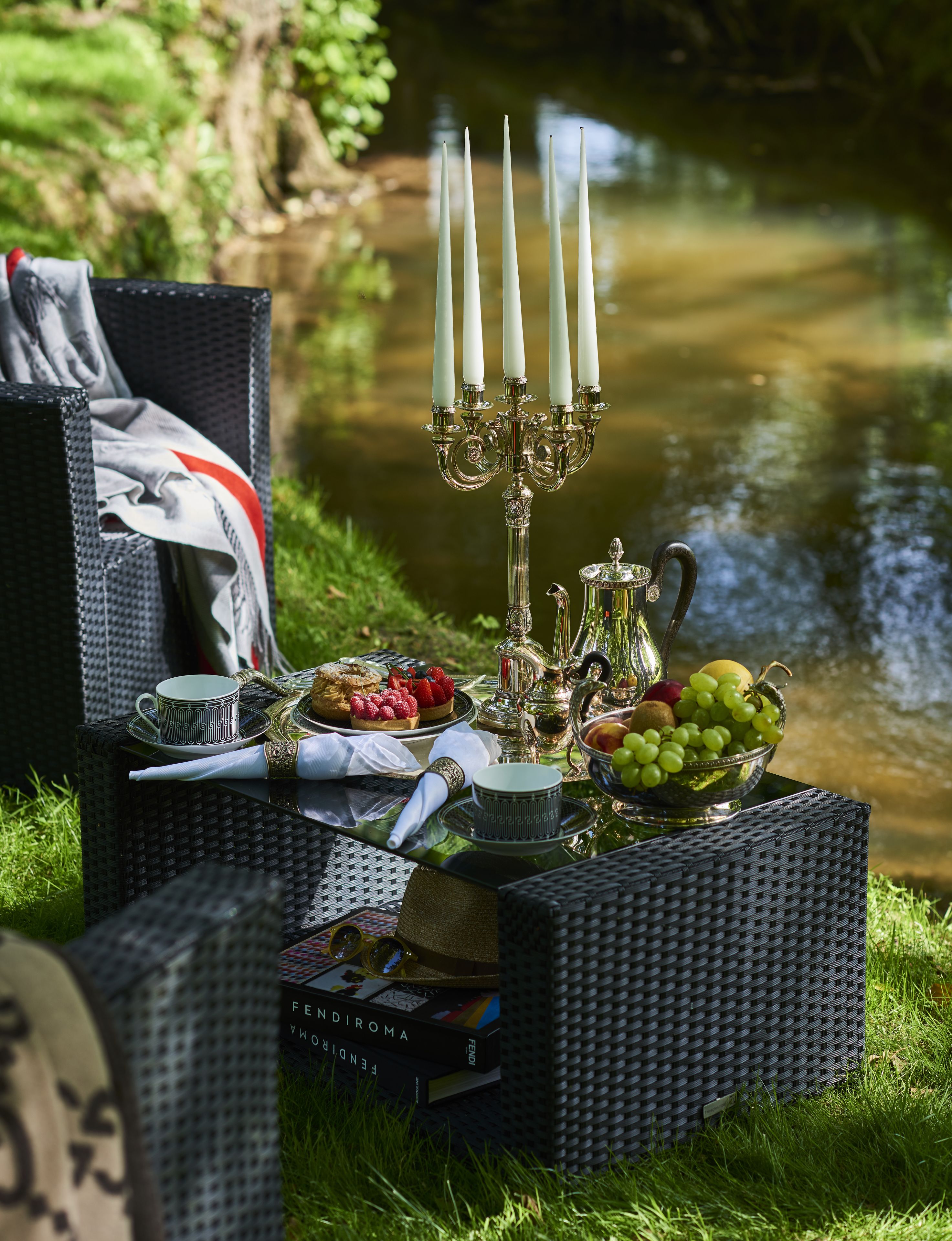 A picnic setup with a wicker table, a candelabra, a fruit bowl, and a book titled "FENDI ROMA" on a grassy area near a body of water.