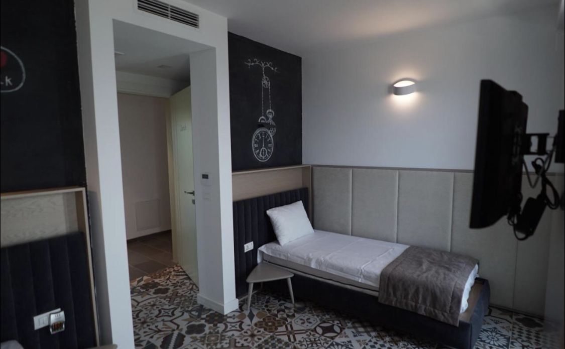 A small, modern room with a single bed, a wall-mounted clock, and a patterned floor.