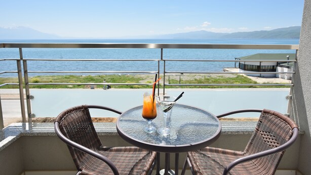 A balcony with wicker chairs and a table, overlooking a body of water and distant mountains. - Vila Europa (Struga)