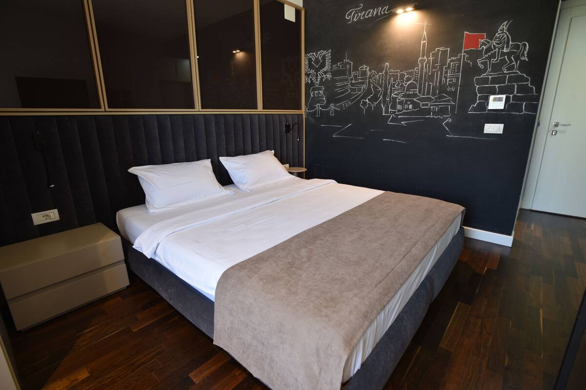 Un lit bien fait, recouvert d’une couverture beige, dans une chambre moderne dont un des murs est décoré d’une fresque représentant un paysage urbain.