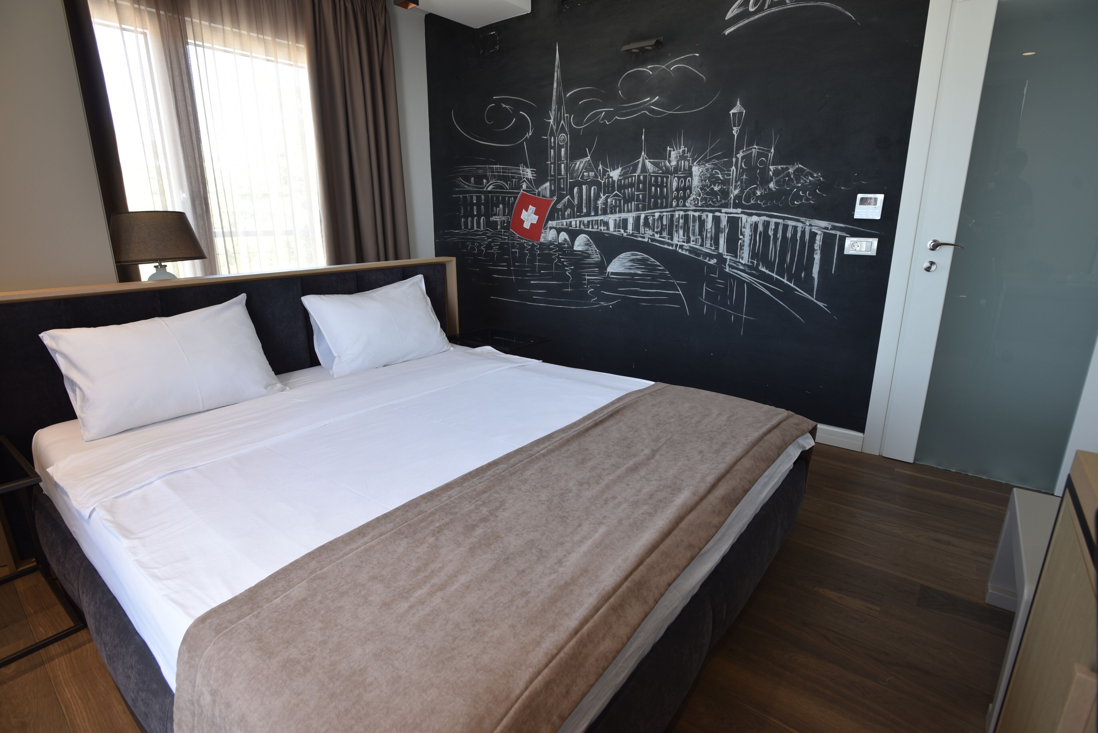 A hotel room with a bed, a nightstand, a lamp, and a wall mural of a cityscape.