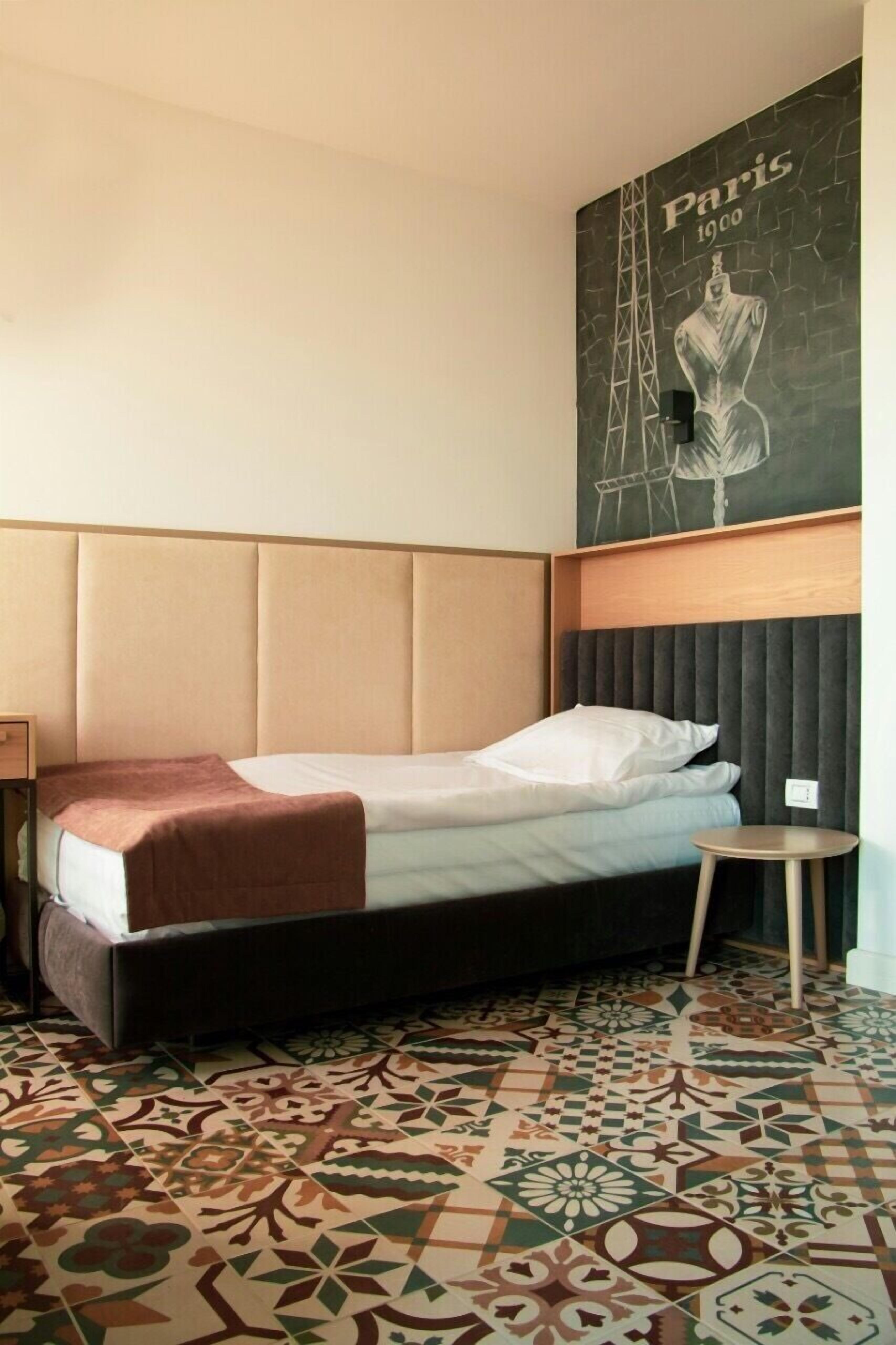 A hotel room with a bed, a small round table, a decorative wall mural, and patterned floor tiles.