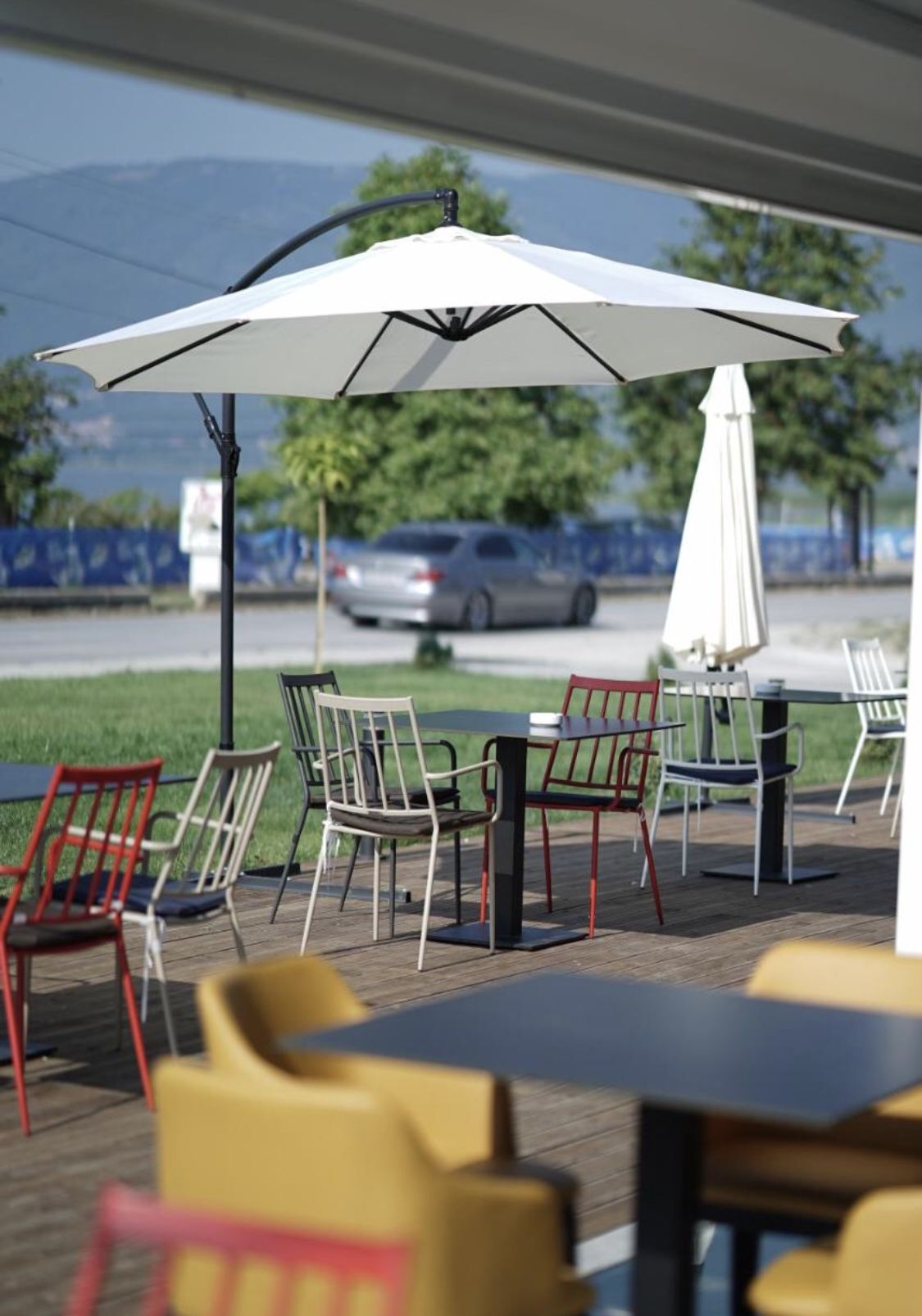An outdoor seating area with tables and chairs, umbrellas, and a view of the street and greenery.