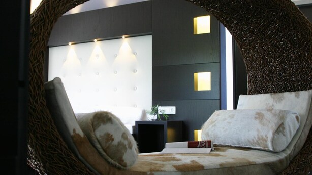 A circular woven chair with a cushioned seat and backrest, a bedside table with a plant, and a wall with three illuminated panels. - Hotel zur Promenade (Donauwoerth)