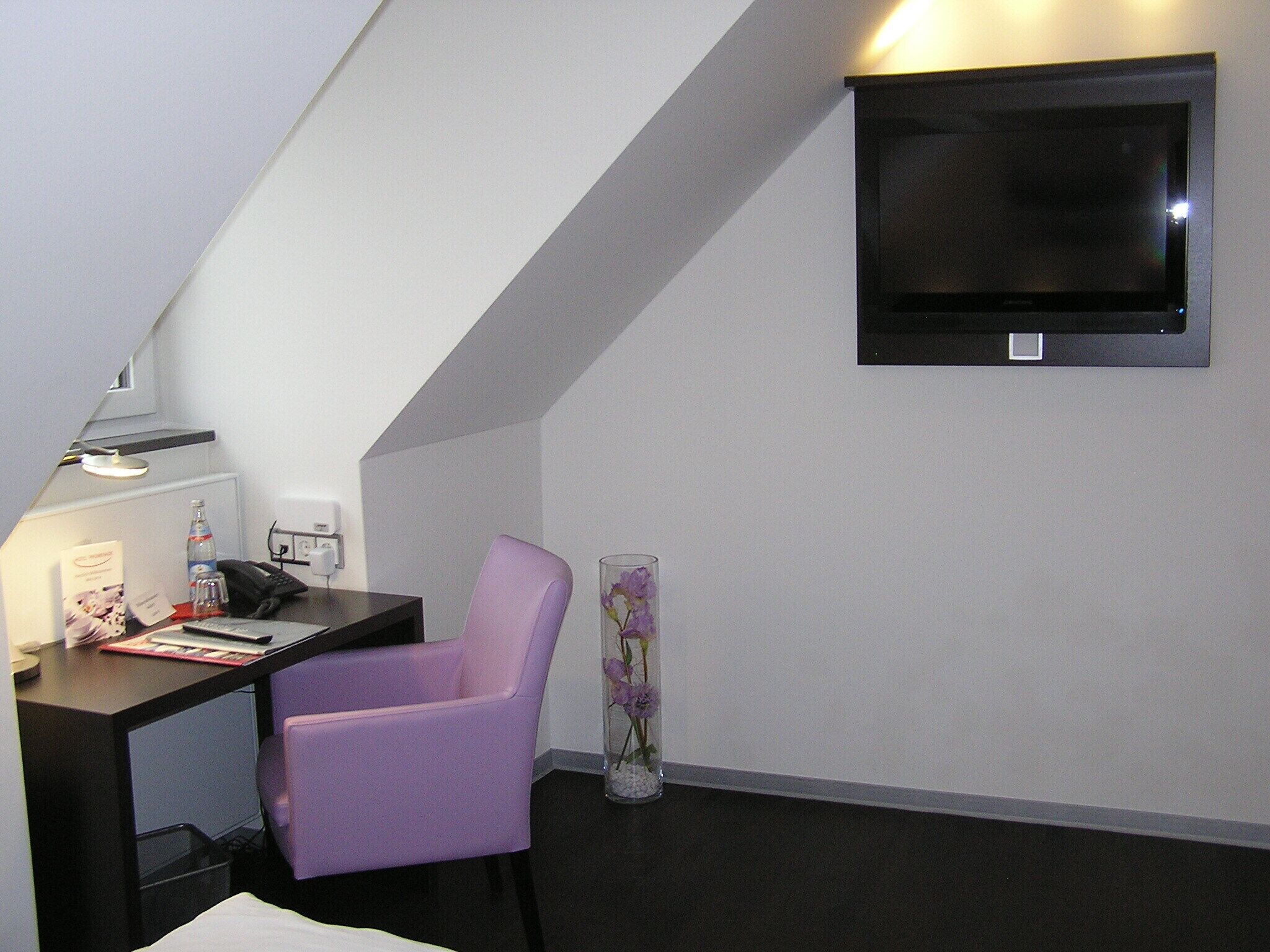 A small, modern room with a desk, a purple chair, a TV mounted on the wall, and a vase with flowers.