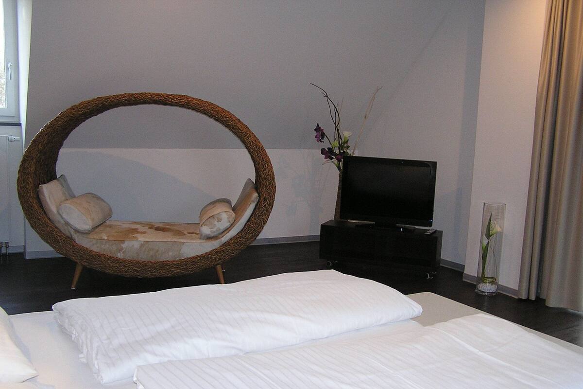 A bedroom with a large bed, a wicker chair, a television, and a vase with flowers.