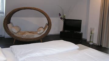 Une chambre avec un grand lit, un fauteuil en osier, une télévision et un vase rempli de fleurs.