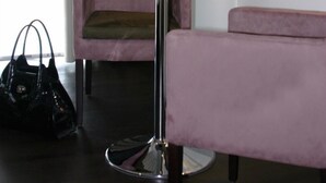 A hotel room with a TV mounted on the wall, a small round table with a tray of items, a pink chair, and a bench. - Hotel zur Promenade (Donauwoerth)
