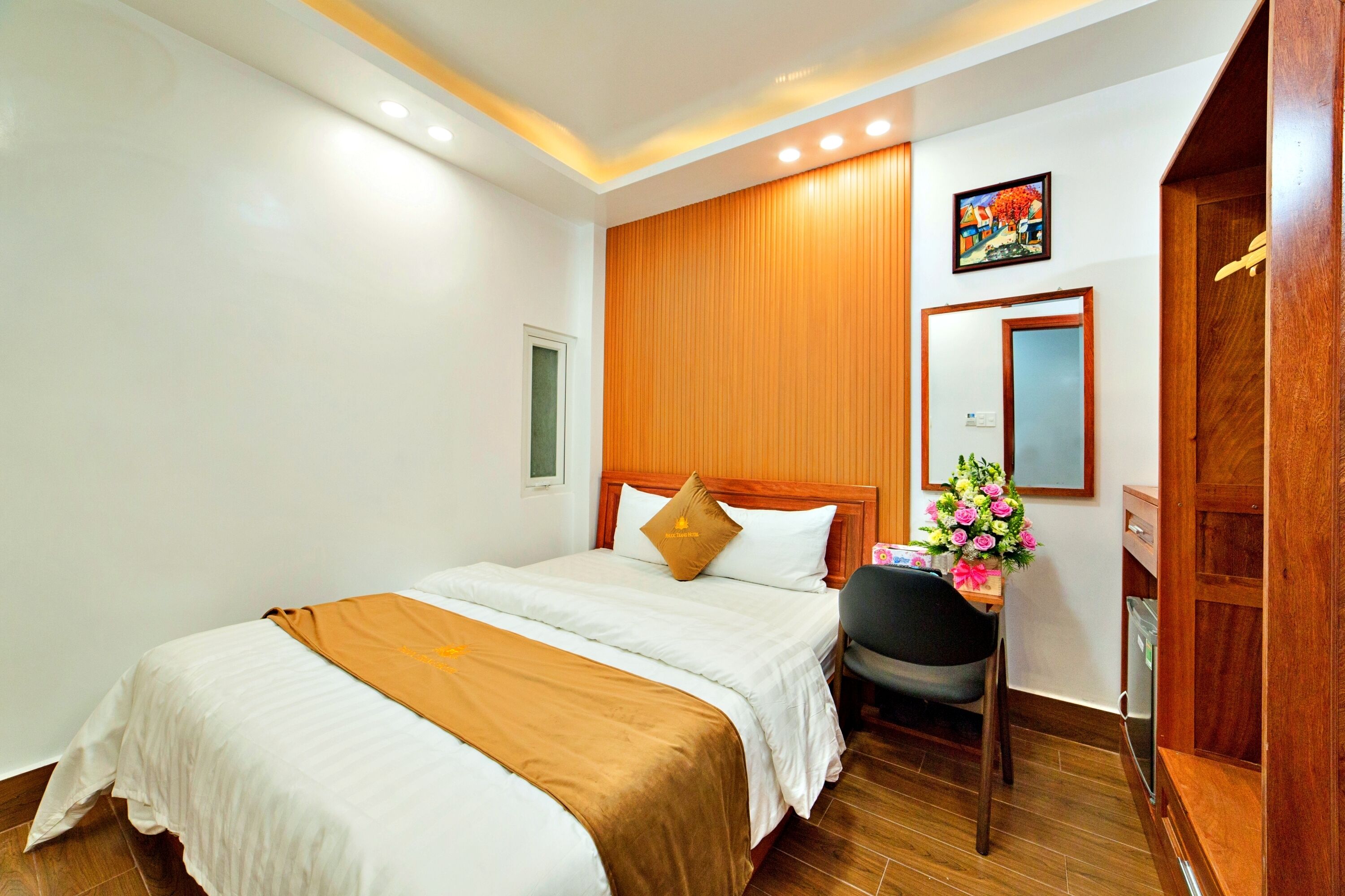 A hotel room with a large bed, a wooden headboard, a desk with a chair, and a small table with a vase of flowers.