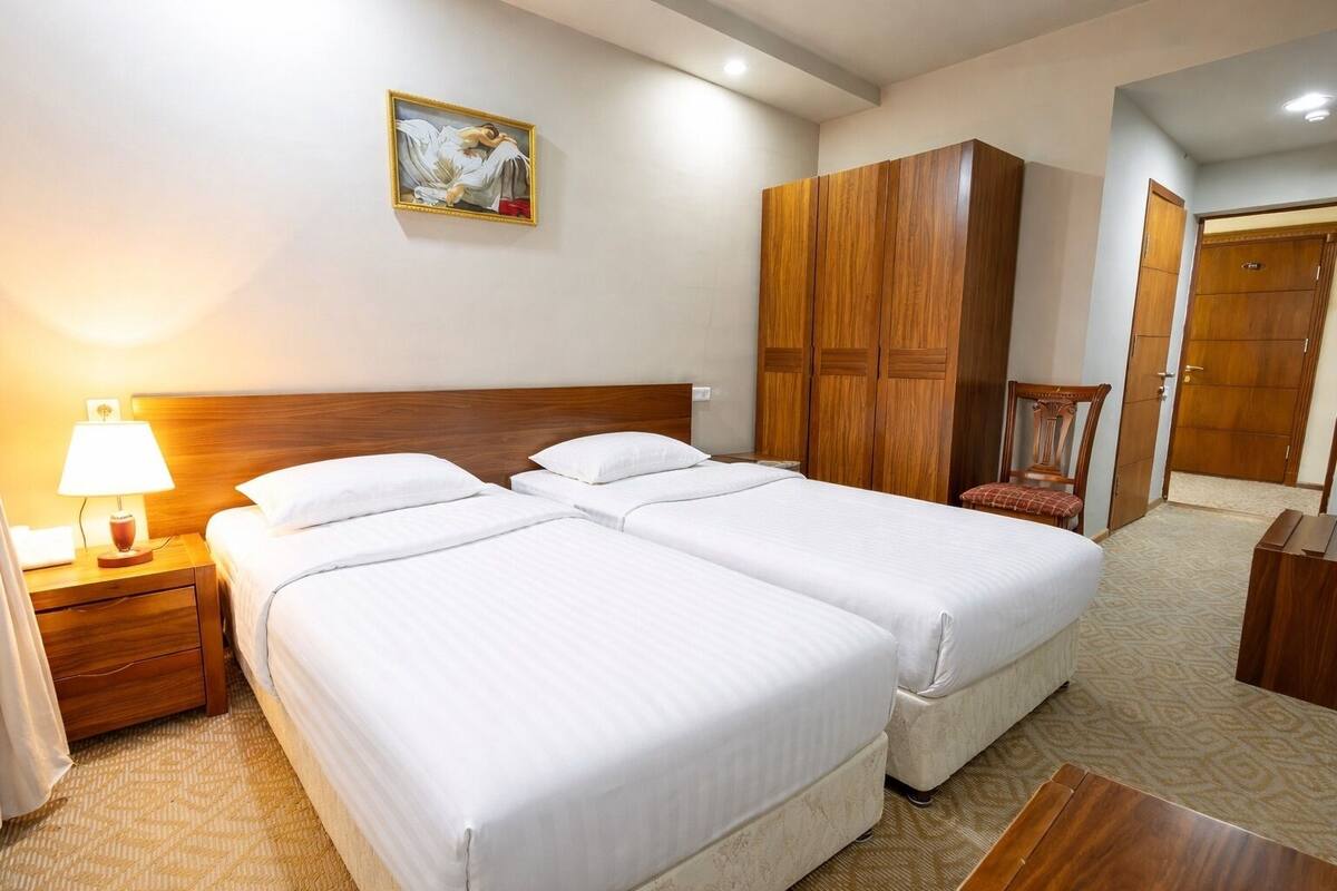 A hotel room with two beds, a wooden wardrobe, a nightstand with a lamp, and a painting on the wall.