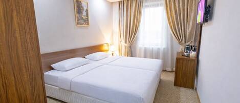 A hotel room with a large bed, a bedside table with a lamp, a TV mounted on the wall, and a window with curtains.