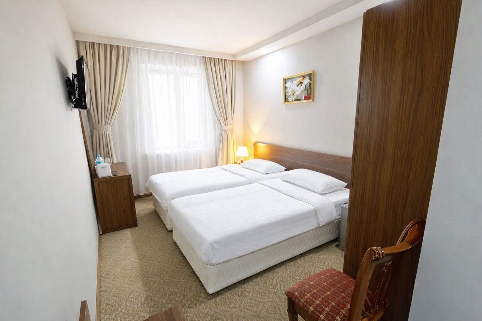 A hotel room with two beds, a desk, a chair, a TV, and a painting on the wall.