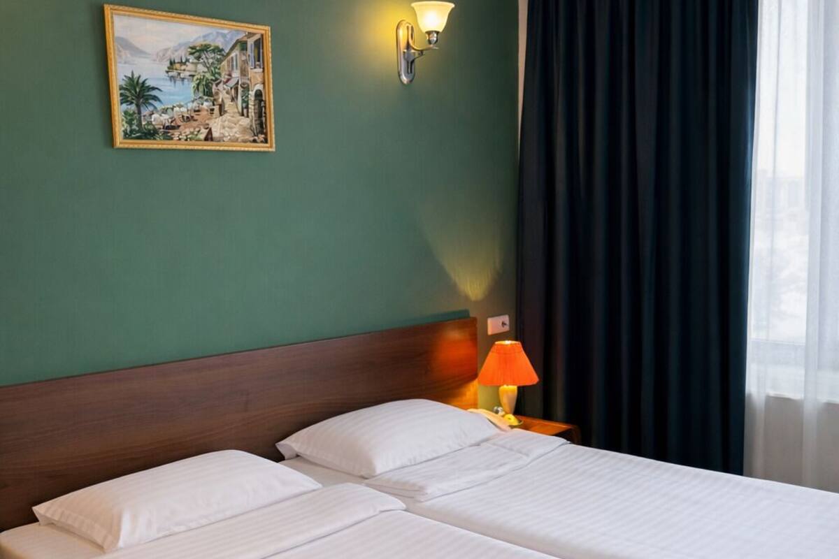 A hotel room with two beds, a nightstand, a lamp, a painting, and a window with curtains.