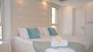 A neatly made bed with white linens and decorative pillows, a glass-topped bedside table, and a window with natural light. - Renovated White 3BD next to Beach - Pool (Eilat)