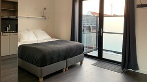 Un dormitorio moderno con una cama, un armario, una mesita y una puerta corrediza de vidrio a un balcón.