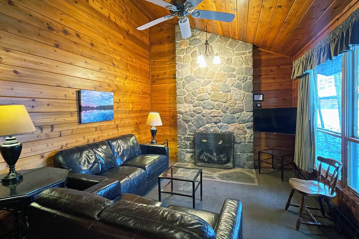 A cozy cabin interior with wooden walls, a stone fireplace, leather sofas, and a television.