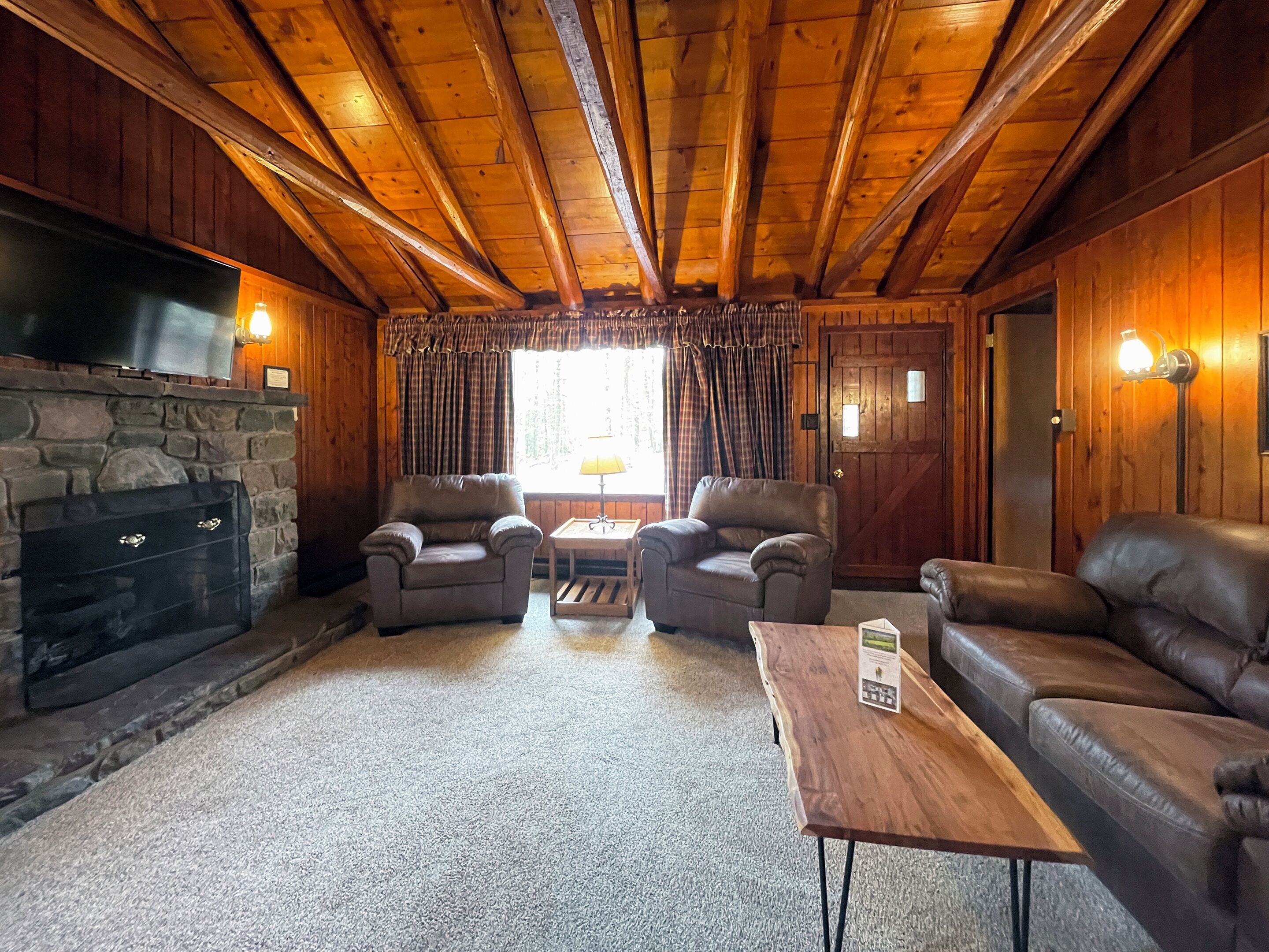A cozy cabin interior with a stone fireplace, two leather armchairs, a wooden coffee table, and a television mounted on the wall.