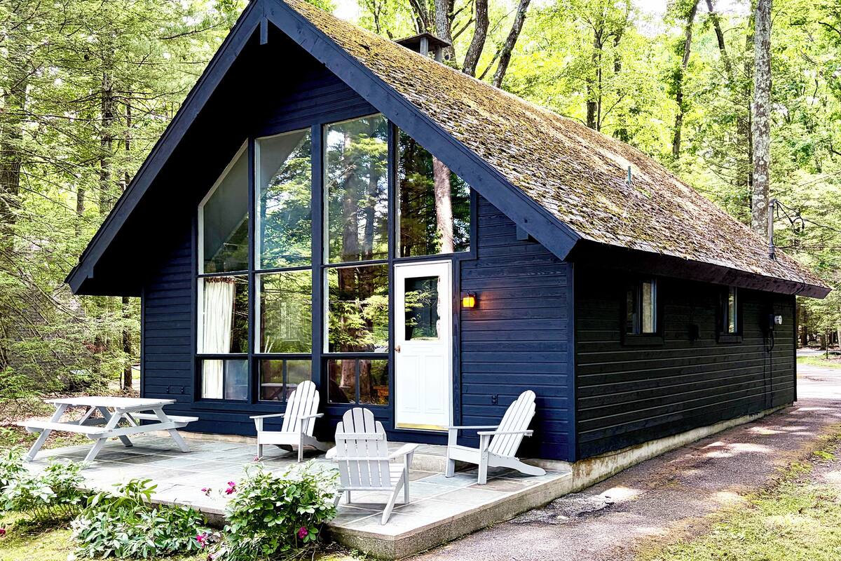 A cabin with a large glass window, a thatched roof, and a picnic table with chairs.