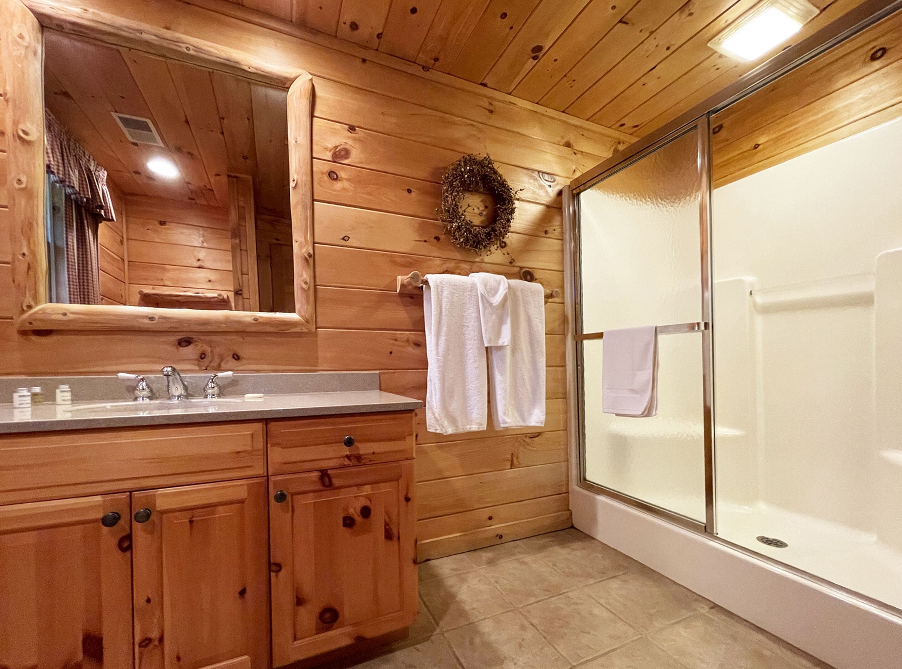 Baño de cabaña de madera con un espejo grande, un lavabo con grifos, cerramiento de ducha de vidrio y un perchero para toallas.