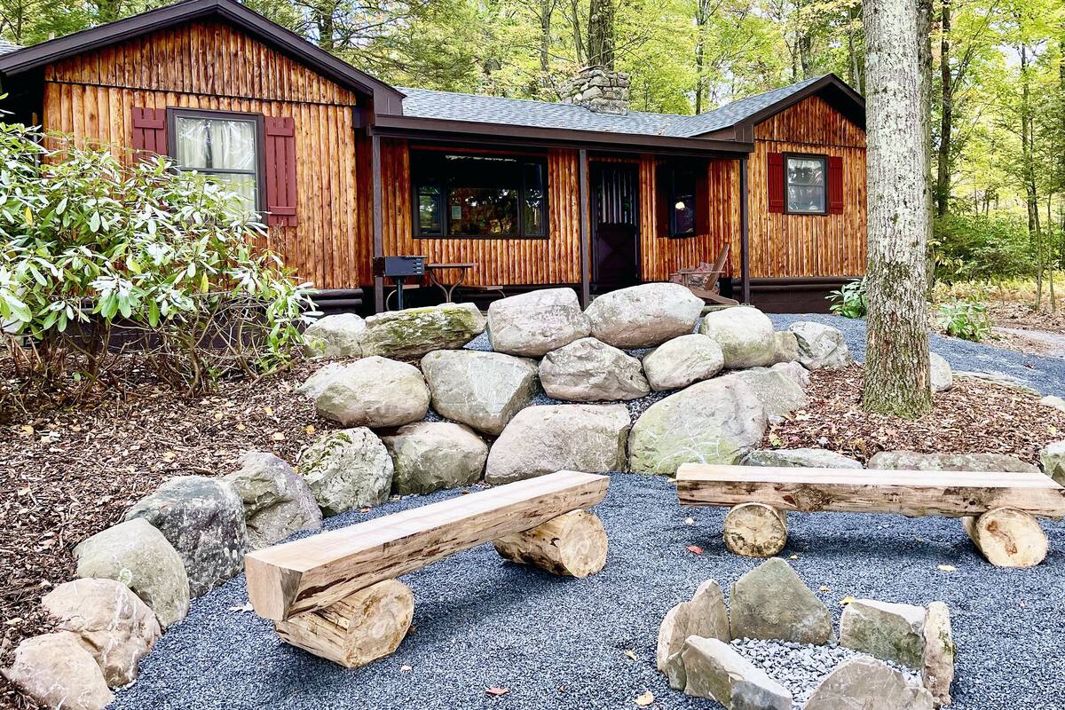 A rustic cabin with a stone wall entrance, wooden benches, and a gravel path.