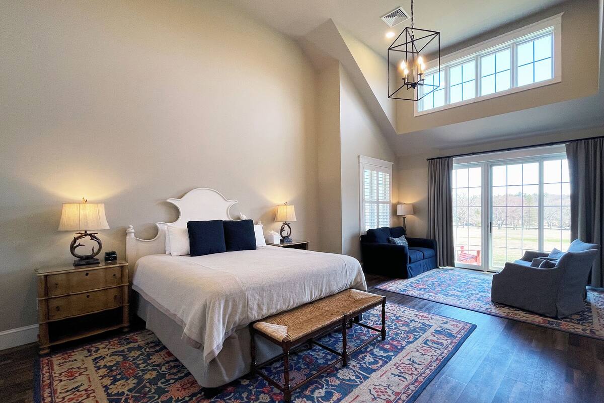 A spacious bedroom with a large bed, a bench, a sofa, and a chair. There are two bedside tables with lamps, a large window, and a ceiling light fixture.