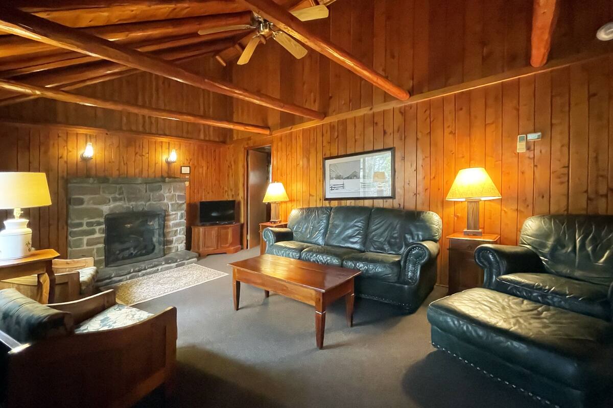 A cozy log cabin interior with a stone fireplace, leather sofas, wooden coffee table, and a ceiling fan.