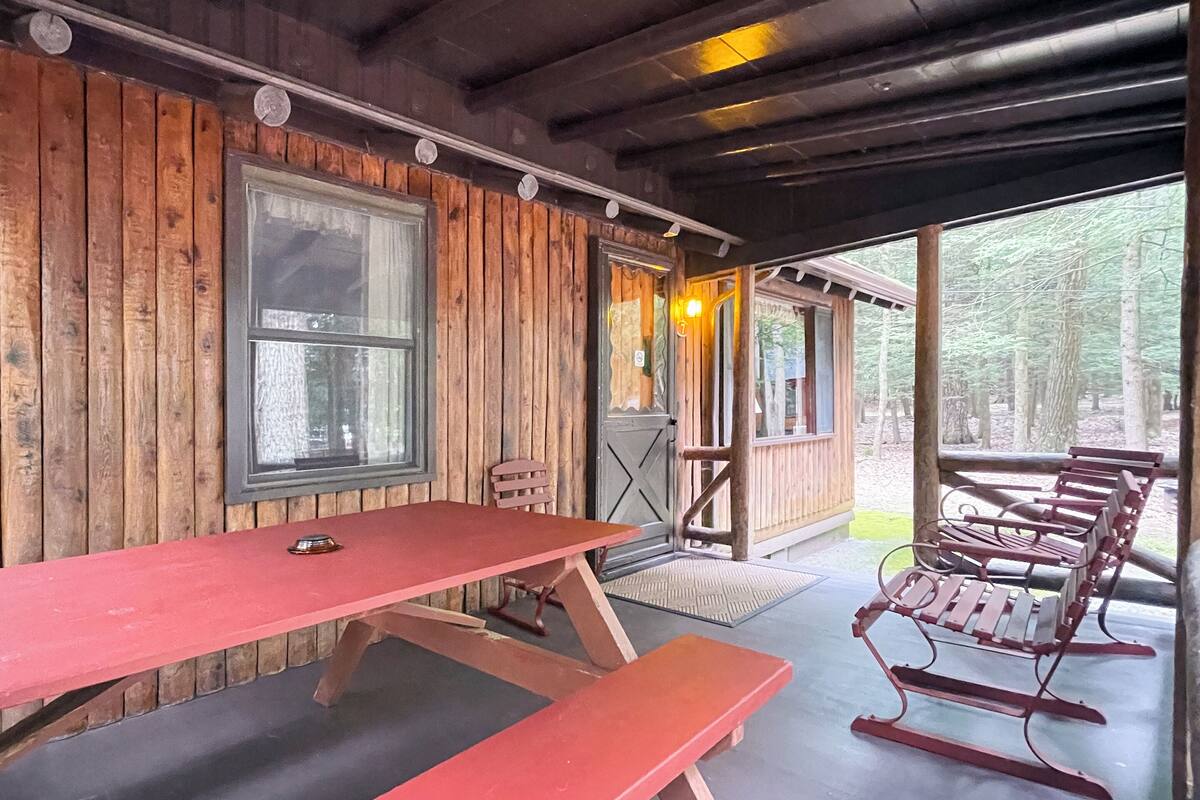 A wooden cabin with a covered porch, a table, benches, and a view of the forest.