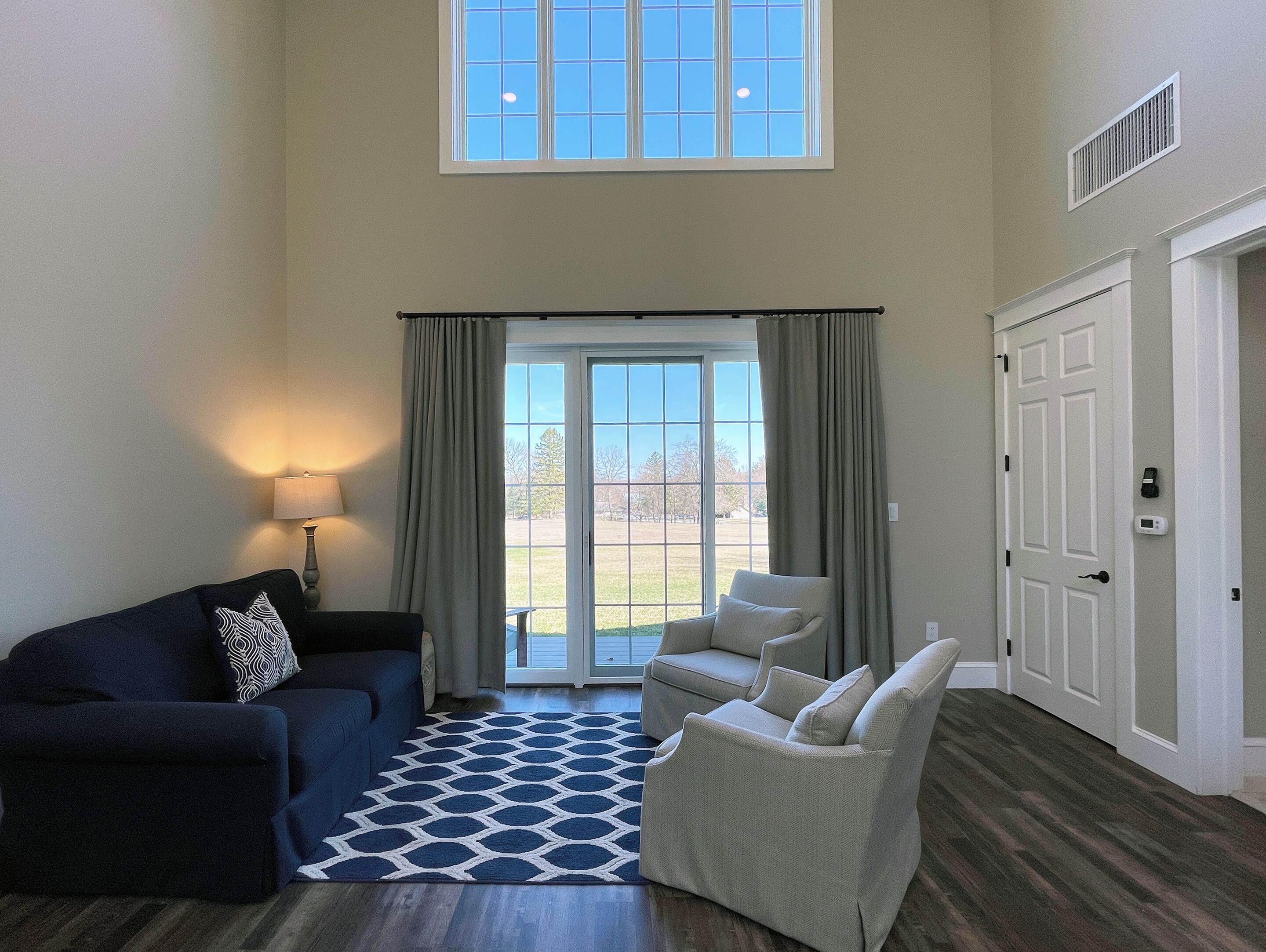 A spacious living room with a dark blue sofa, a beige armchair, and a patterned rug. A large window with curtains allows natural light to enter.