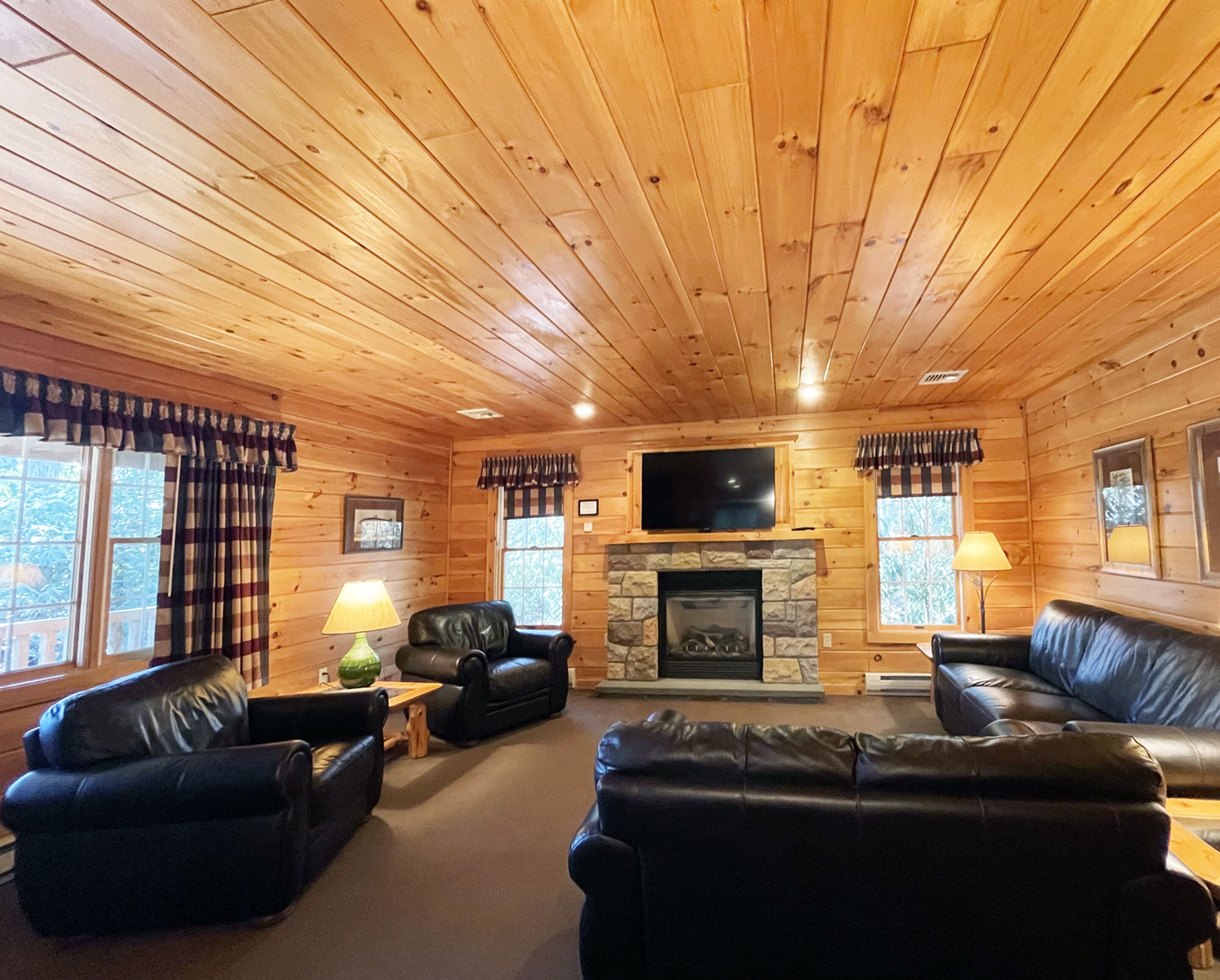 A log cabin interior with a stone fireplace, leather sofas, and a television mounted above it.