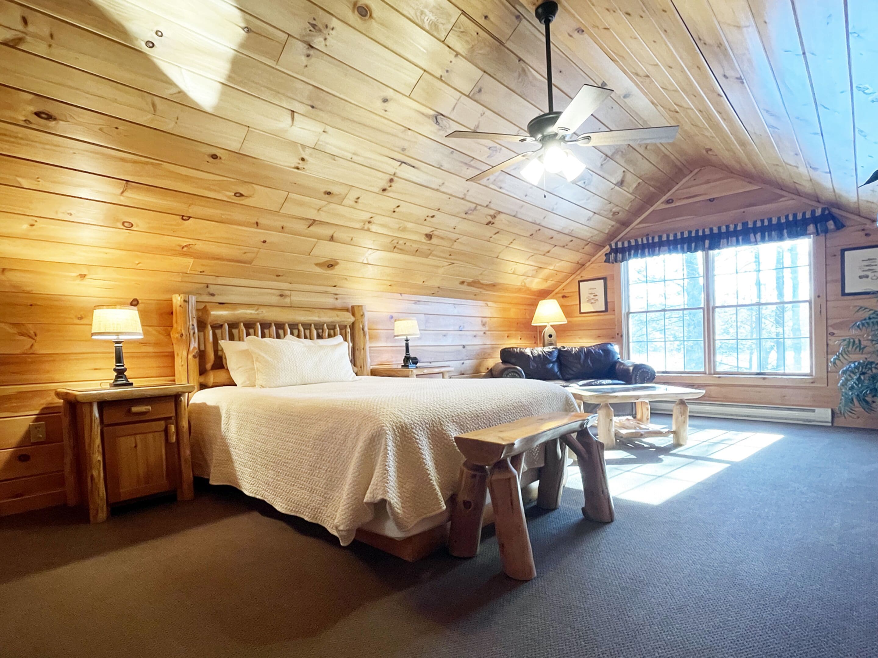 Un dormitorio acogedor tipo cabaña de madera con techo de madera, una cama con cabecera, dos mesitas de noche con lámparas, un banco y un ventanal con vistas a árboles.
