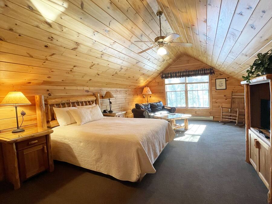 A cozy log cabin bedroom with a large bed, two bedside tables with lamps, a rocking chair, a television, and a window with a view of trees.