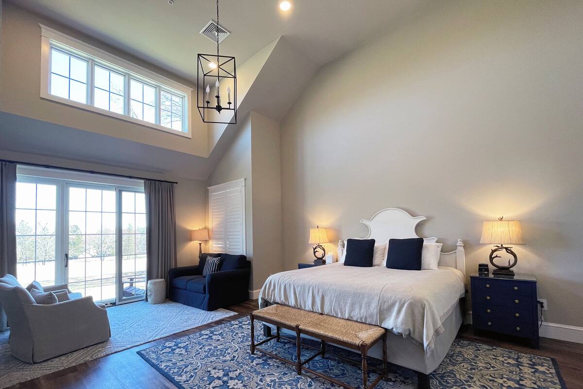 A spacious bedroom with a large bed, a bench, a chair, and a desk. There are two windows and a chandelier.