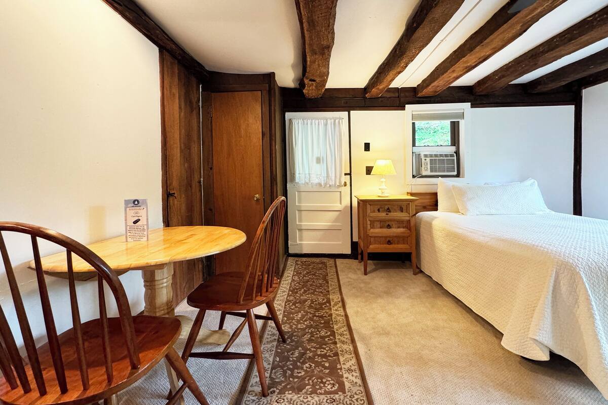 A cozy room with a wooden table and chairs, a bed with white bedding, a nightstand with a lamp, and a window with curtains.