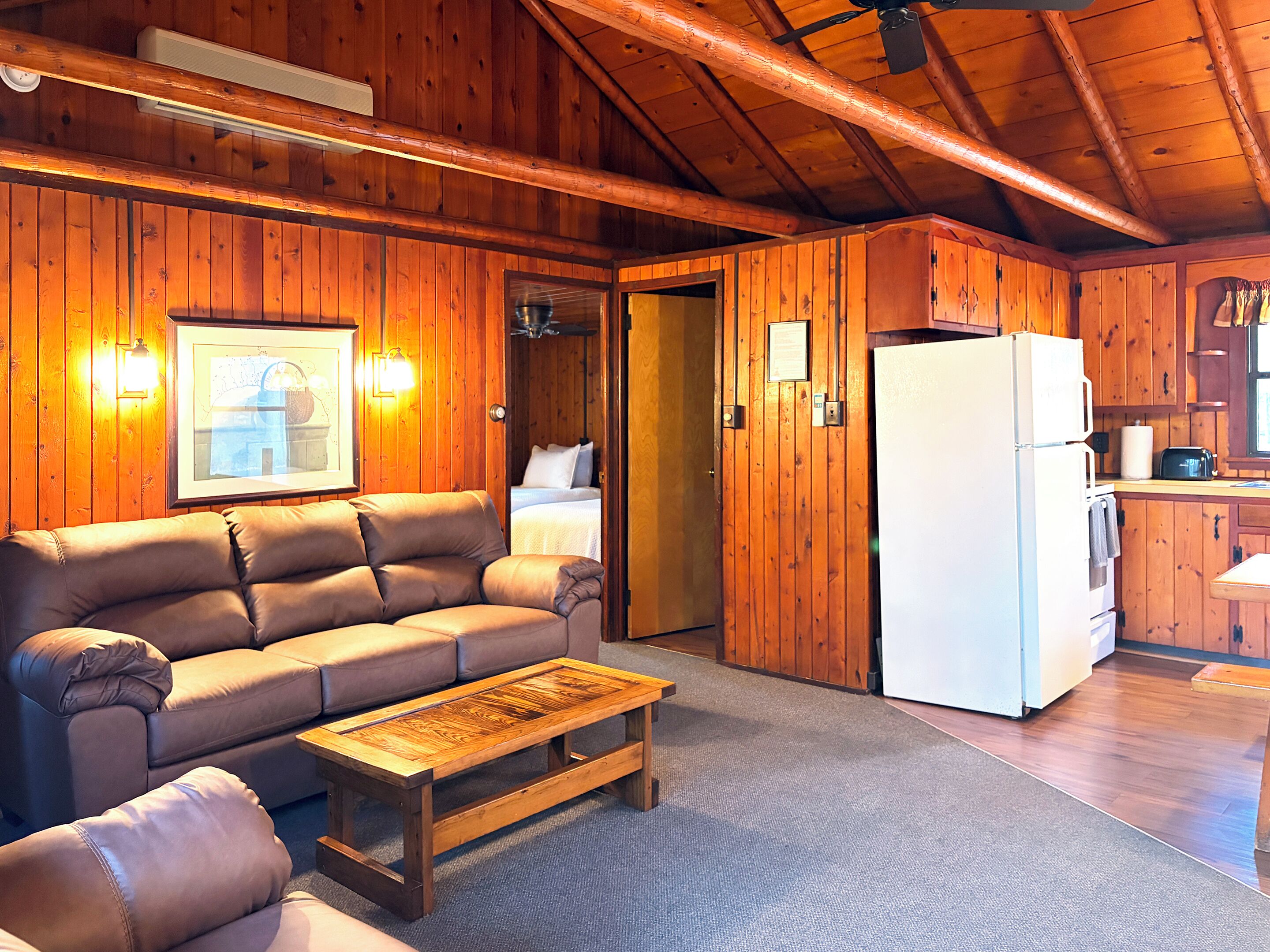 A wooden cabin interior with a sofa, a wooden coffee table, a kitchen area, and a framed picture on the wall.