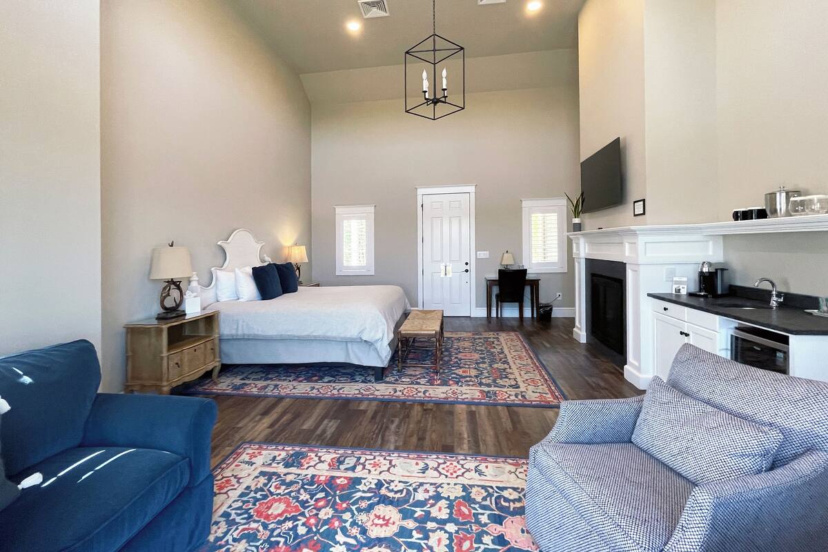A spacious bedroom with a large bed, a sitting area with a sofa and armchair, a fireplace, and a television mounted on the wall.